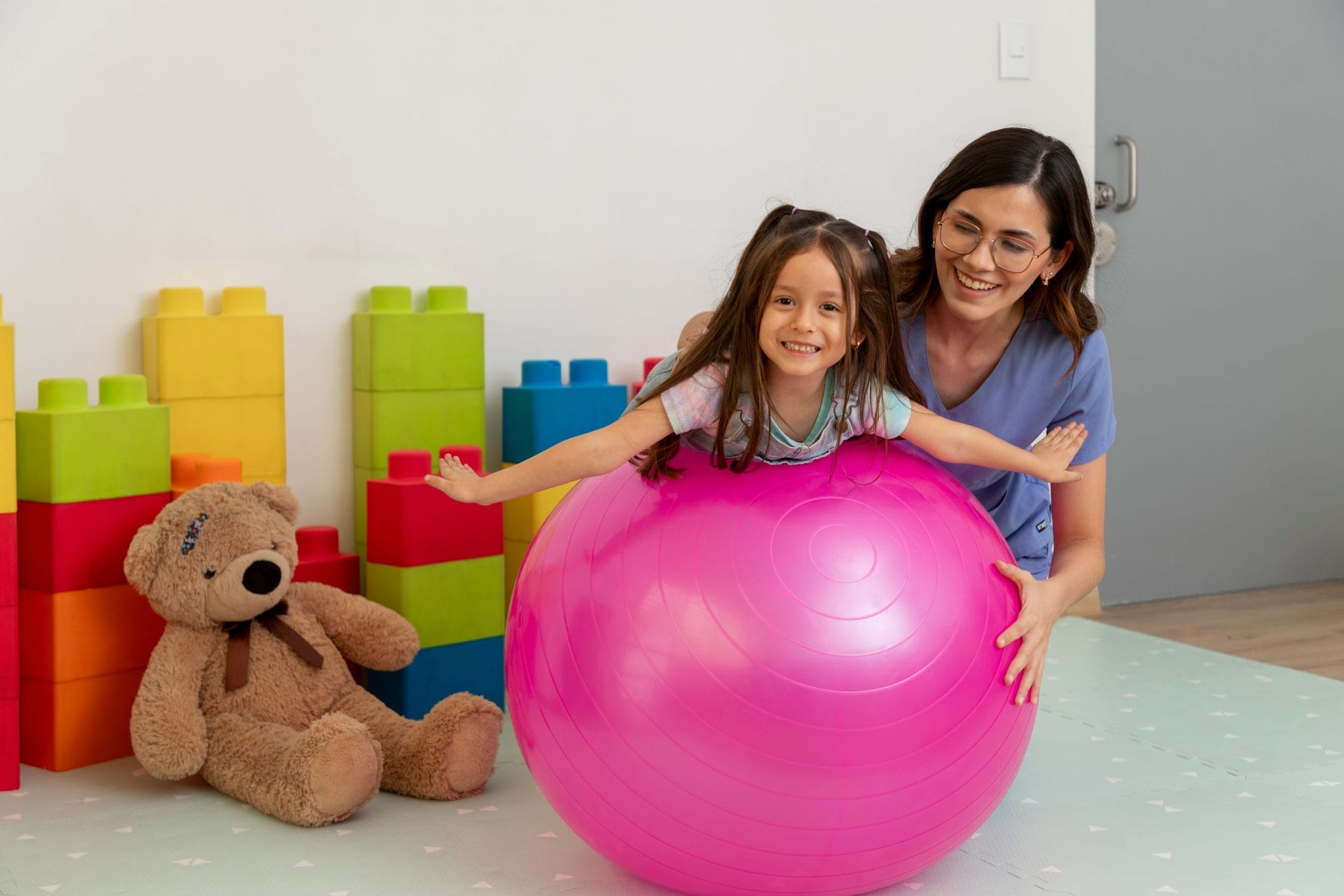 Two girls sitting on the floor, playing with a large pink exercise ball.