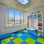 Pediatric room with multicolored foam floor tiles, wall shelving, and large window.