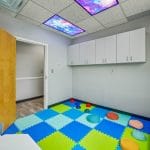Open pediatric room with foam tile floor, white walls, and wooden door.