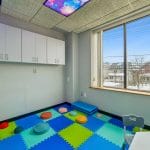 Therapy room with colorful foam flooring, corner shelving, and large window view.