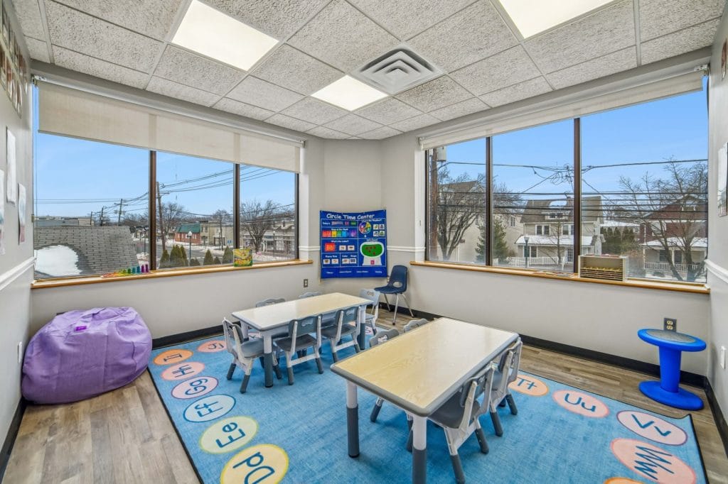 Classroom-style therapy space with child-sized tables, bright rug, and city view through wide windows.