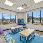 Classroom-style therapy space with child-sized tables, bright rug, and city view through wide windows.