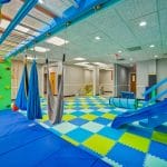 Wide view of sensory gym featuring obstacle elements, swings, and bright foam mats.