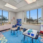 Pediatric therapy room with small table and chairs, colorful rug, whiteboard, and large window with natural light.