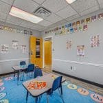Therapy room corner with educational posters, blue flooring, and child-sized seating near window.