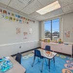 Pediatric room with blue rug, small table, wall art, and wide window overlooking neighborhood.