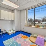 Therapy area with soft seating, colorful floor mat, and large window providing natural daylight.